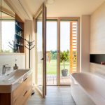 Moderná kúpeľňa s vaňou a umývadlom, dreveným nábytkom a vstupom do záhrady cez sklenené dvere.
