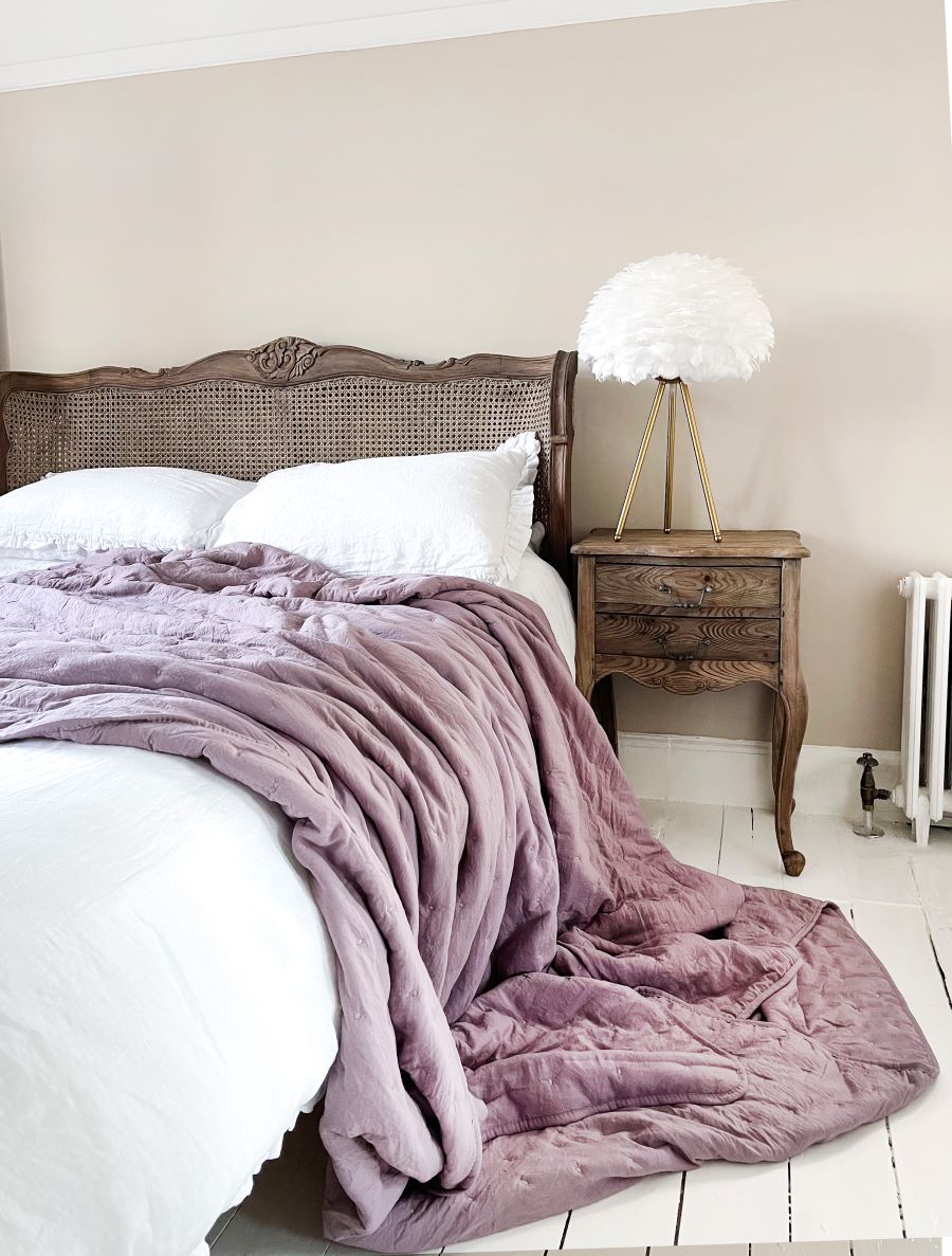 Časť postele orgovánovou dekou, bielymi obliečkami a čelom i nočným stolíkom z tmavého dreva.