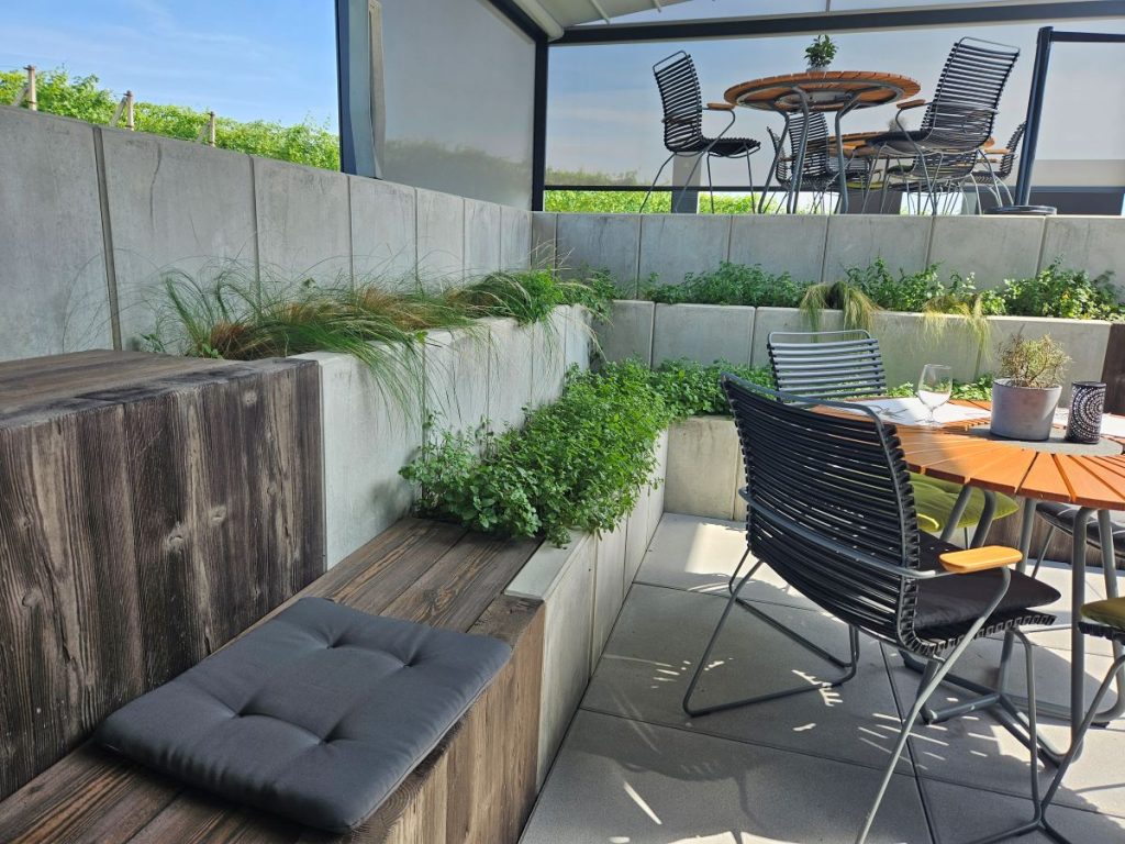 Členitý terén využitý na vytvorenie súkromia pri vonkajšom posedení na terase vinárstva v kombinácii pohľadového betónu, imitácii dreva a čiernym kovom vonkajšieho nábytku.