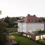 Pohľad na historickú vilu na pražských Vinohradoch pri súmraku so svietiacimi oknami a záhradou.
