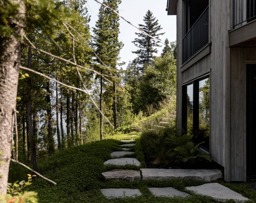 Záhrada so skalným chodníkom vedúcim popri modernom dome, obklopená vysokými stromami a hustou vegetáciou.