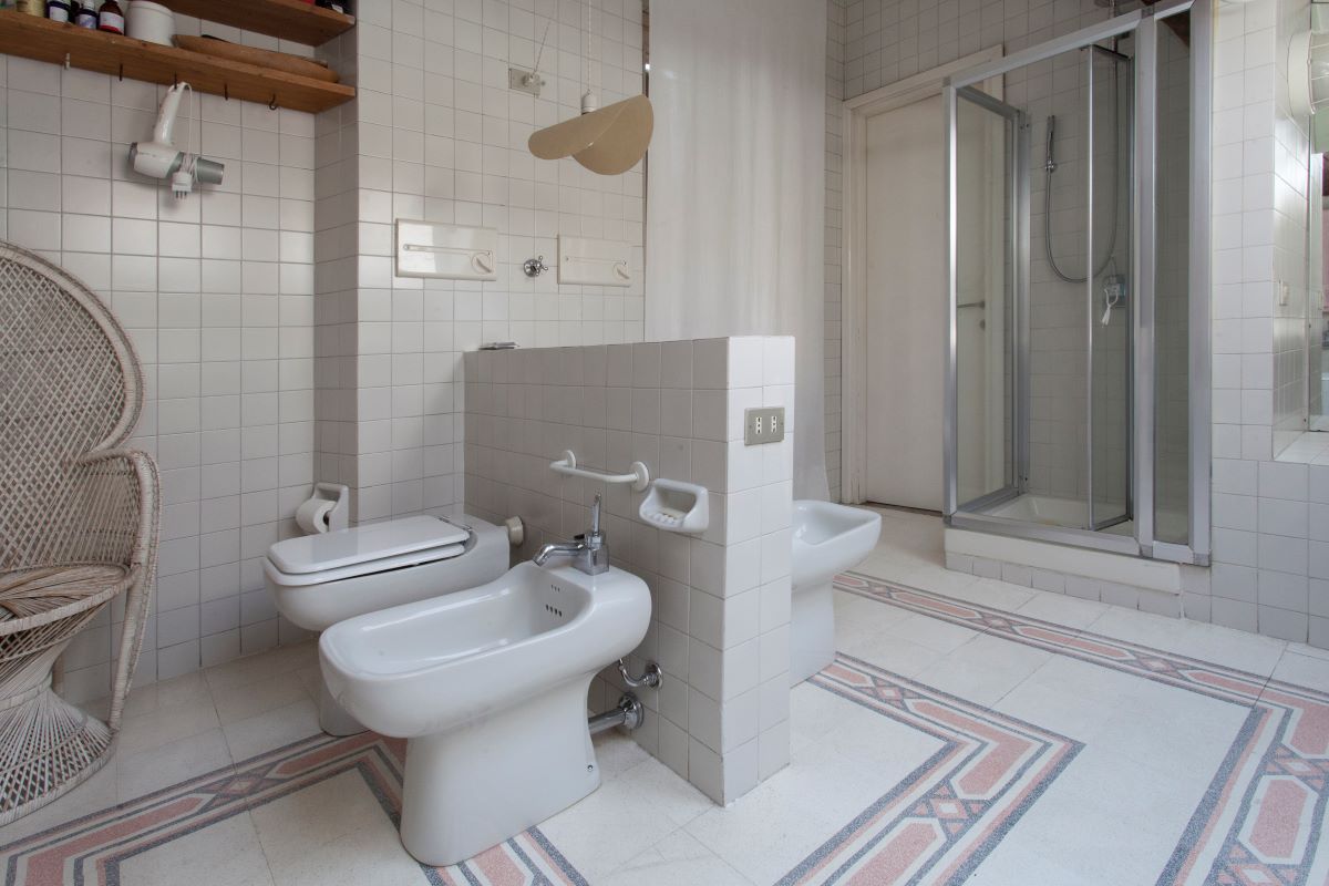 Priestranná kúpeľňa s bielym obložením s toaletou, bidetom, sprchovacím kútom a ratanovým kreslom.