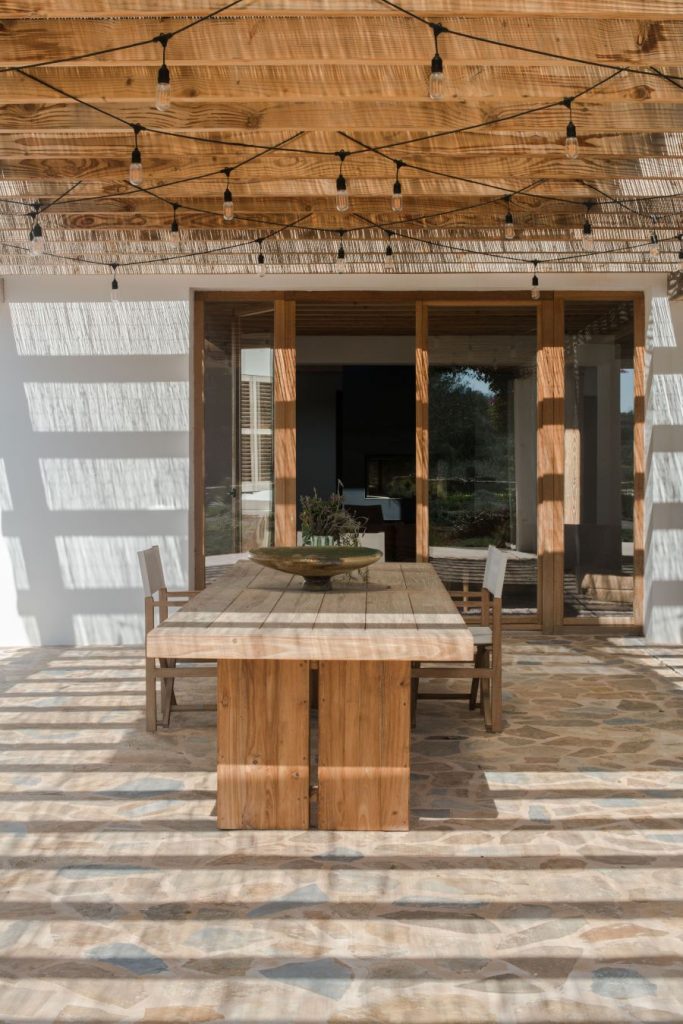 Pohľad na terasu pod drevenou pergolou s masívnym dreveným stolom so stoličkami a vstupom do interiéru domu.