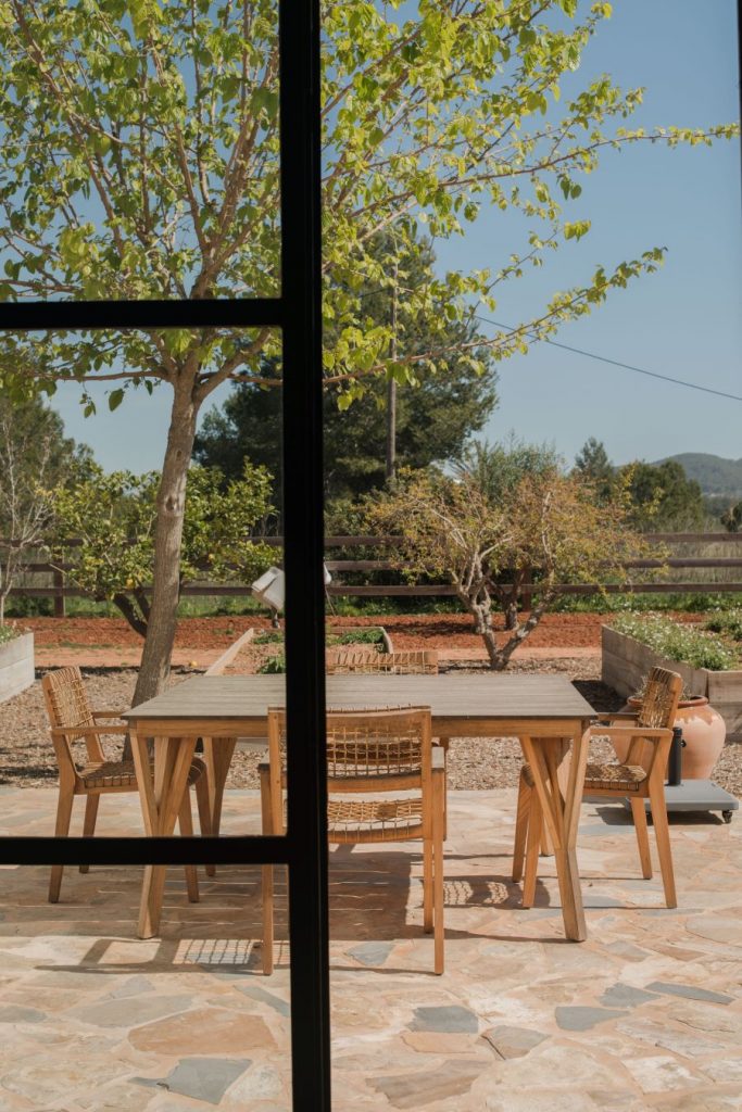 Pohľad cez sklenené dvere na vonkajšiu terasu s dreveným stolom a stoličkami, obklopenú zeleňou.