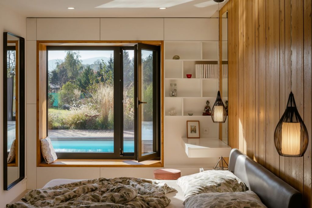 Moderne zariadená spálňa s posteľou, oknom do záhrady so záhonmi a bazénom a otvorenými policami s dekoráciami.