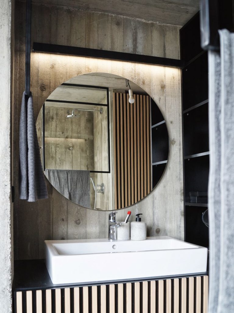 Minimalistická kúpeľňa s dreveným obložením, veľkým okrúhlym zrkadlom a moderným umývadlom.
