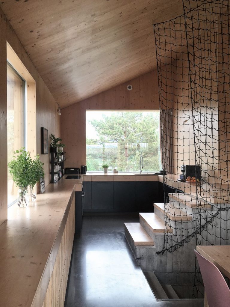 Svetlá kuchyňa s dreveným obložením a veľkými oknami pri schodisku so sieťou, vedúcom do inej časti domu.