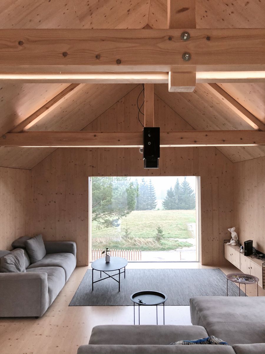 Obývacia izba s vysokým dreveným stropom, veľkým oknom, nadčasovými sedačkami a minimalistickým nábytkom.