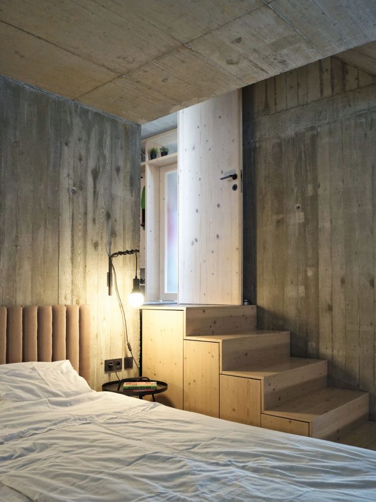 Časť spálne s posteľnou s čalúneným čelom, priznaným betónom na stenách a drevenými schodmi do vyššej úrovne.