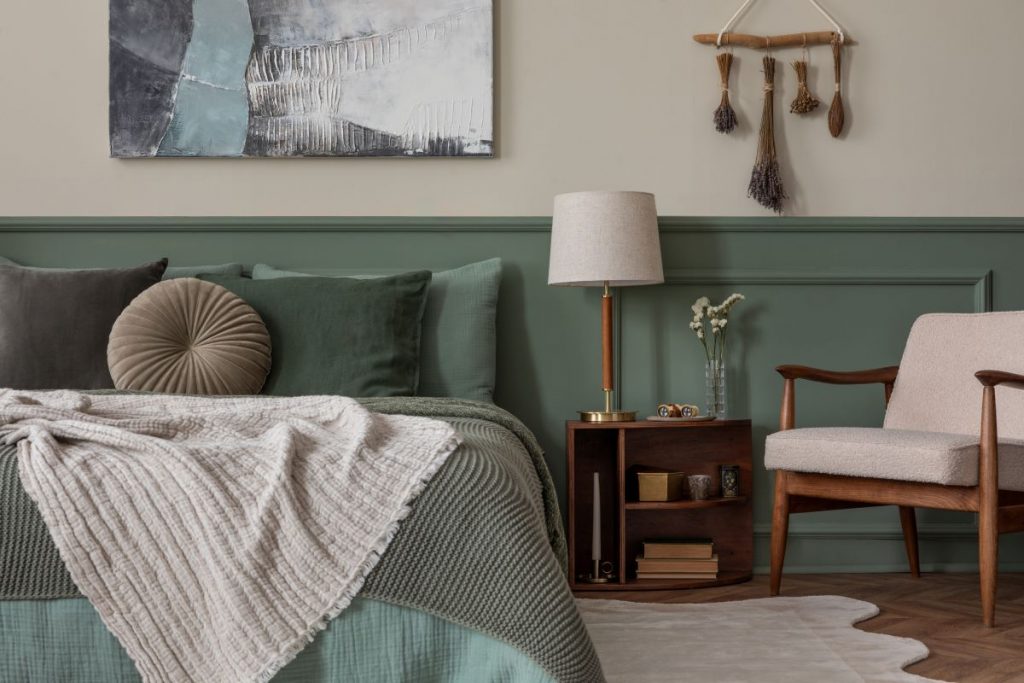Spálňa s posteľnou bielizňou a stenou v šalviovej zelenej farbe, svetlým kreslom s drevenou konštrukciou a nadčasovými doplnkami.