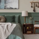 Spálňa s posteľnou bielizňou a stenou v šalviovej zelenej farbe, svetlým kreslom s drevenou konštrukciou a nadčasovými doplnkami.
