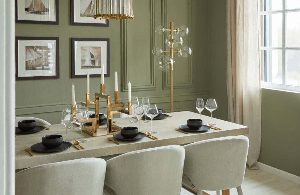 Elegantná jedáleň so svetlým stolom, čalúnenými stoličkami zlato-čiernymi doplnkami a stenou v šalviovej zelenej.