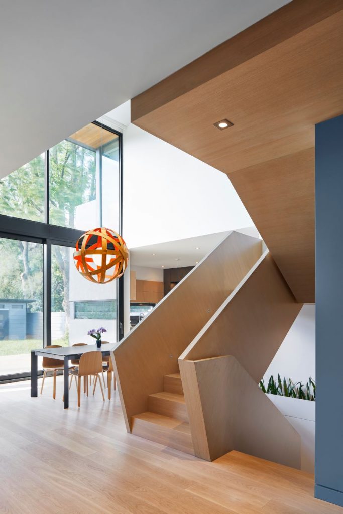 Drevené schodisko v interiéri novostavby pri jedálni s veľkými oknami a dizajnovým osvetlením.