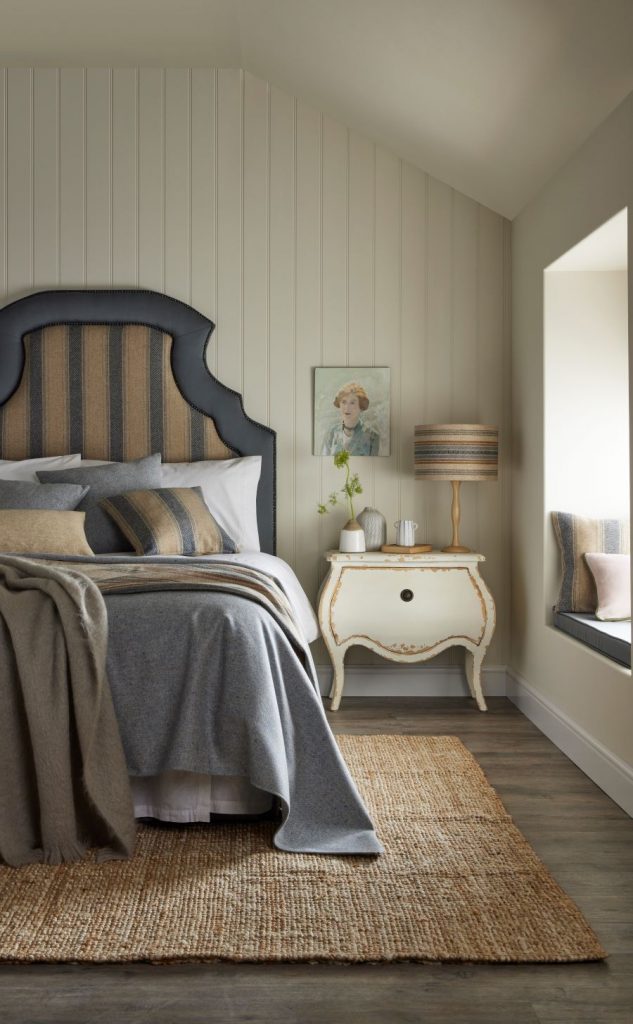 Spálňa zariadená vo vidieckom štýle s posteľou s vysokým čalúneným čelom a bielym nočným stolíkom so zlatými detailmi vo vintage štýle.