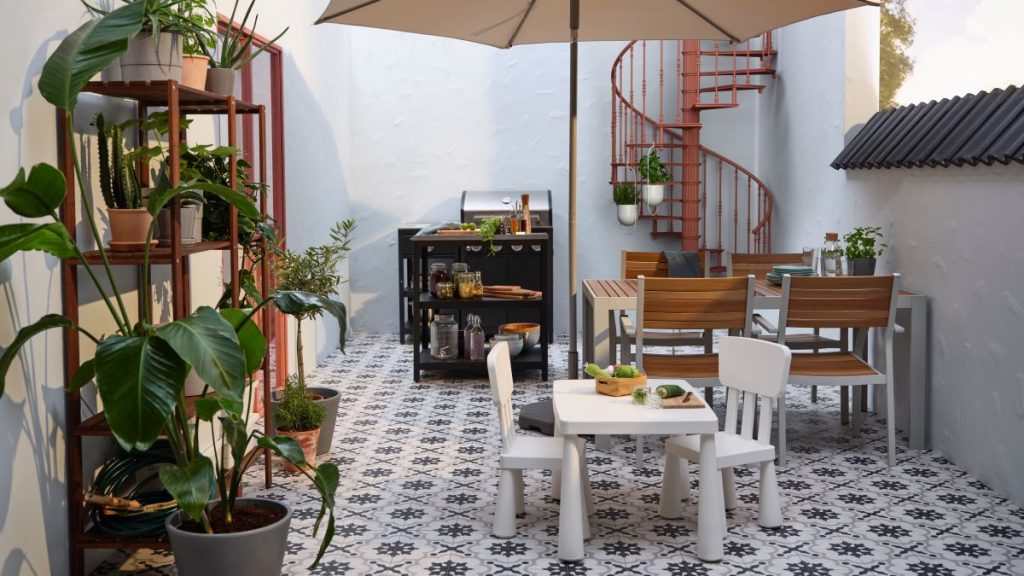 Moderná vonkajšia terasa s dreveným jedálenským stolom, grilom a točitým schodiskom, obklopená bohatou vegetáciou a kvetináčmi.