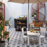 Moderná vonkajšia terasa s dreveným jedálenským stolom, grilom a točitým schodiskom, obklopená bohatou vegetáciou a kvetináčmi.