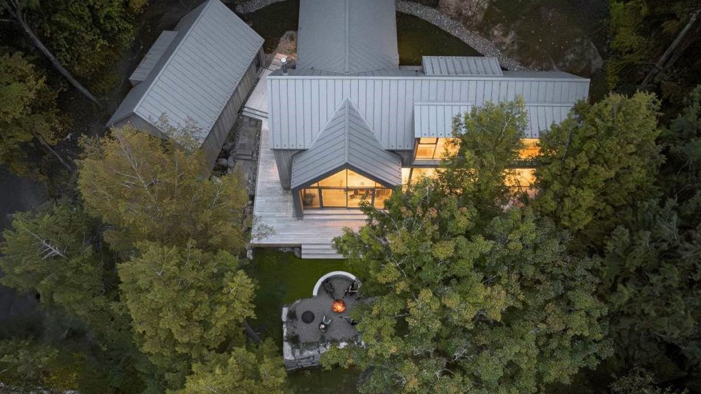 Letecký pohľad na moderný dom s dreveným obložením a ohniskom, zasadený v hustom lesnom prostredí.