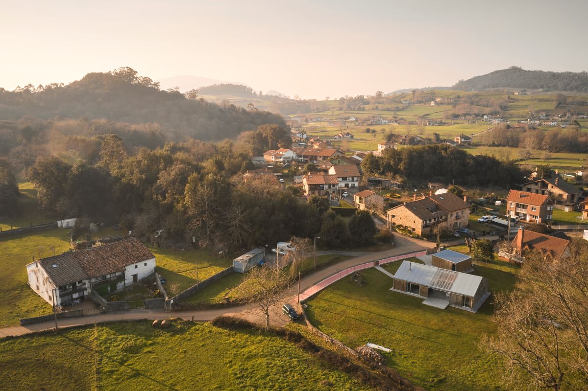 Letecký pohľad na malú vidiecku dedinu s moderným domom, obklopenú lesom a horami.