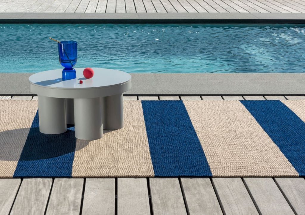 Pruhovaný koberec O deck Electric Blue s nízkym stolom pri bazéne.