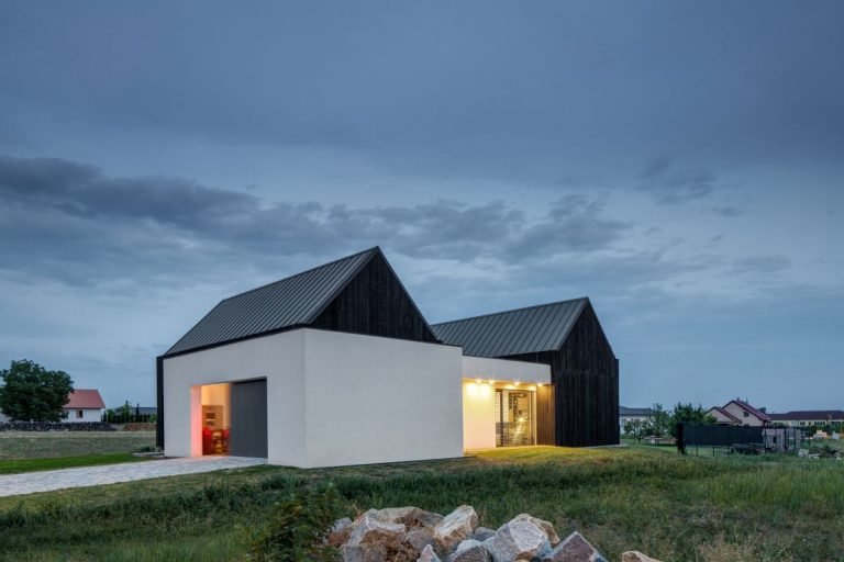 Celkový pohľad na dom za súmraku s dvoma čiernymi strechami, bielou fasádou a osvetlenými oknami.