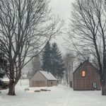 Dve budovy s tradičným tvarom, drevenou a kamennou fasádou a sedlovou strechou v zime za súmraku medzi stromami.