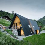 Pohľad na chatu VA House s podsvieteným interiérom a horskou panorámou za súmraku.