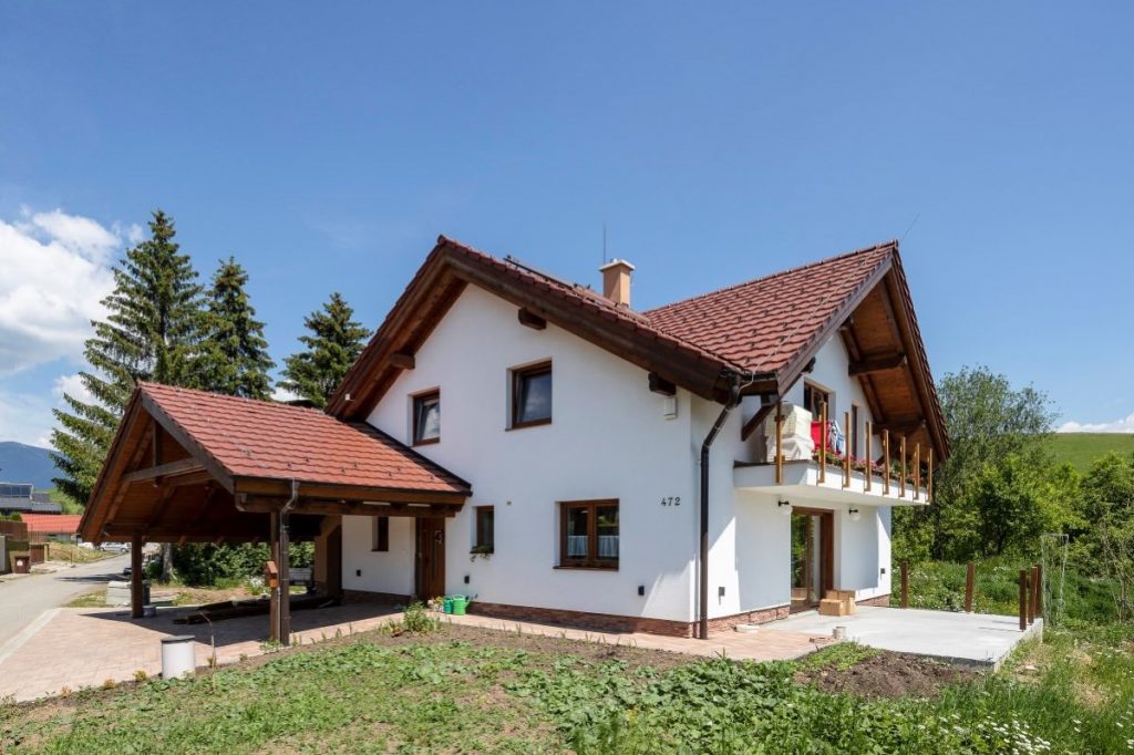 Tradičný vidiecky dom s bielou fasádou, prestrešeným parkovacím miestom a červenou tehlovou strechou.