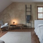 Útulná spálňa s rustikálnou omietkou, tradičné štvorcové okno, minimalistické zariadenie, nízky drevený stolík, útulné kreslo.