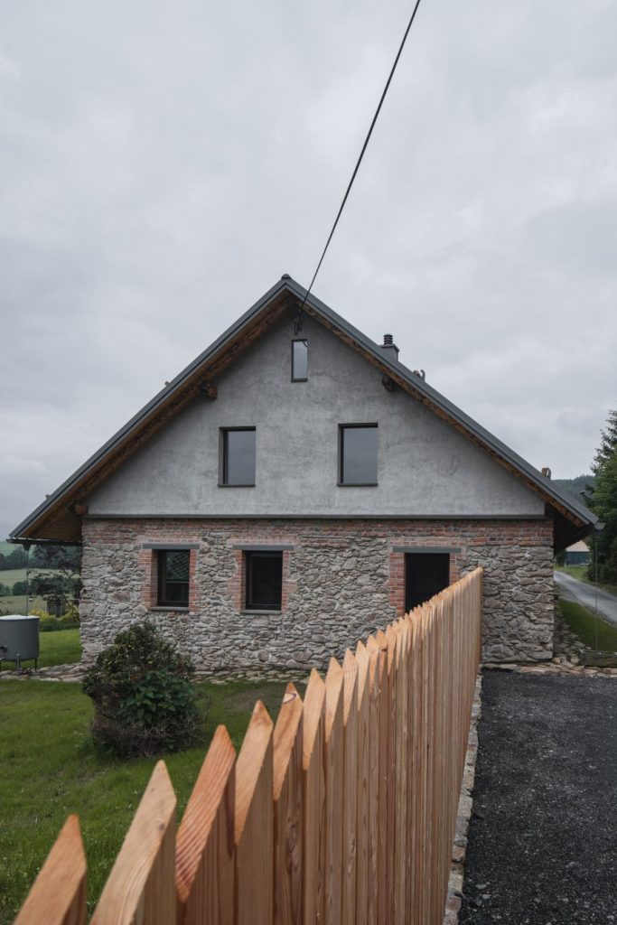 Zadná strana chalupy s kamenným základom a dreveným plotom.