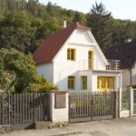Moderný biely dom so žltými oknami a balkónom, obklopený zeleňou a susednými budovami.