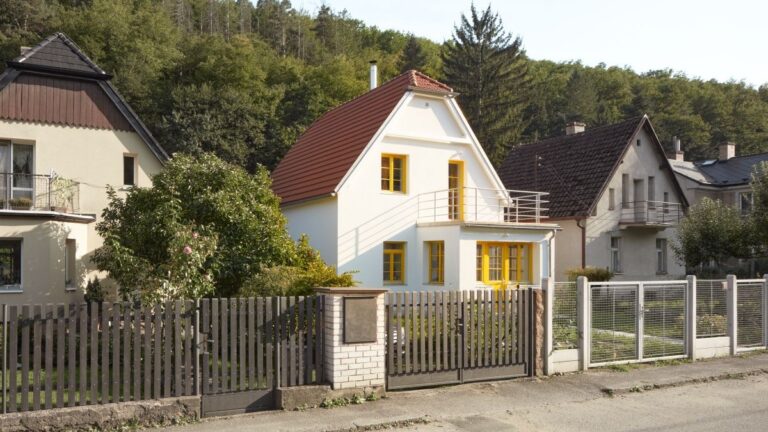 Moderný biely dom so žltými oknami a balkónom, obklopený zeleňou a susednými budovami.