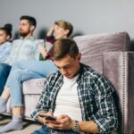 Mladý hostiteľ pri sedačke pozerá do mobilu a nevenuje sa návšteve.