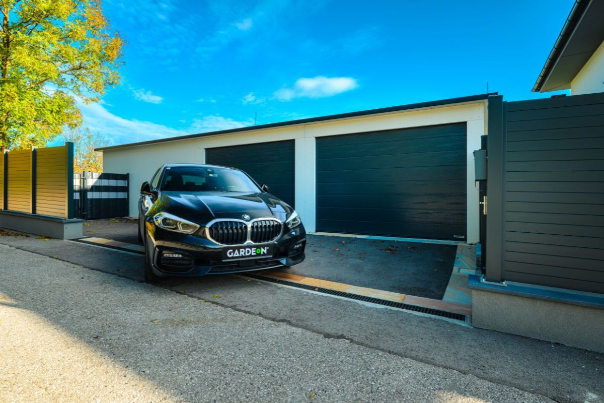 Moderná čierna garážová brána so senzorom, čierne BMW parkuje pred garážou pri záhrade.