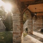 Kamenná pergola s dreveným stropom a oblúkovými stĺpmi, s výhľadom na drevenú chalupu.