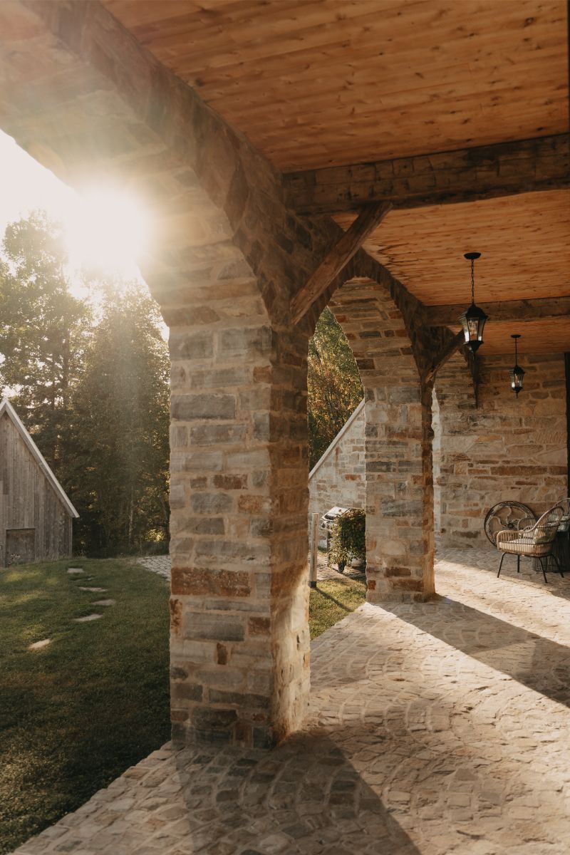 Kamenná pergola s dreveným stropom a oblúkovými stĺpmi, s výhľadom na drevenú chalupu.