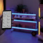 Ruka držiaca smartfón s aplikáciou pre ovládanie smart osvetlenia s modrými LED pásmi v pozadí.