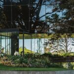 Detail presklenej fasády moderného domu s reflexným čiernym sklom a obklopením zeleňou, s betónovou rampou a rastlinami.