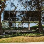 Fasáda moderného domu s čiernym zrkadlovým sklom a vlnitou betónovou rampou, zasadená do lesného prostredia.