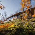 Moderný dom s tmavou fasádou a veľkými oknami, zasadený do svahu v lese s jesennou vegetáciou.