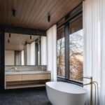 Luxusná kúpeľňa s bielou voľne stojacou vaňou, dreveným stropom a veľkým oknom s výhľadom do prírody.
