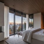 Moderná spálňa s krbom, dreveným stropom a veľkými oknami so svetlými závesmi a výhľadom na jazero.