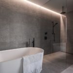 Moderná kúpeľňa v šedej farbe s veľkoformátovým obkladom, bielou vaňou a preskleným sprchovým kútom.