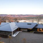 Fasáda moderného domu so šedou plechovou strechou a kameňom, zasadeného do prírody s výhľadom na jazero.