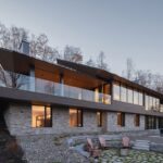 Viacpodlažný moderný dom s kamennou fasádou, drevenými prvkami a terasou s preskleným zábradlím.