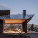 Večerný záber na modernú drevenú terasu s vonkajším sedením a presklenou stenou s plechovou strechou.