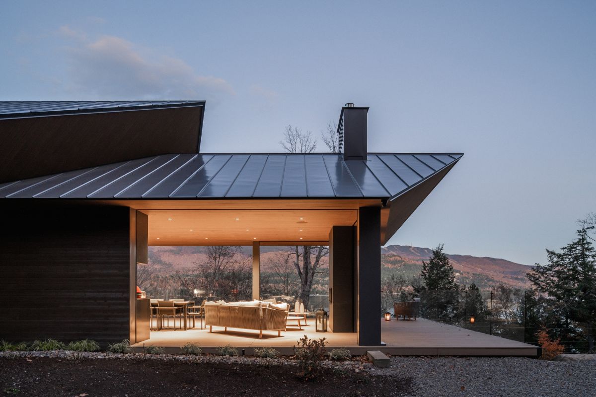 Večerný záber na modernú drevenú terasu s vonkajším sedením a presklenou stenou s plechovou strechou.