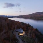 Panoramatický pohľad z vtáčej perspektívy na moderný dom s tmavou strechou na brehu jazera obklopený lesom.