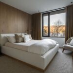 Luxusná spálňa s bielou čalúnenou posteľou, dreveným obkladom na stene a hnedými závesmi s veľkým oknom.