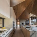 Dlhá moderná kuchyňa s bielymi skrinkami, mramorovými pracovnými doskami a šikmým dreveným stropom