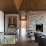 Moderná obývacia izba s kamenným krbom, drevenou podlahou a zaskleným zábradlím vedúcim na poschodie.